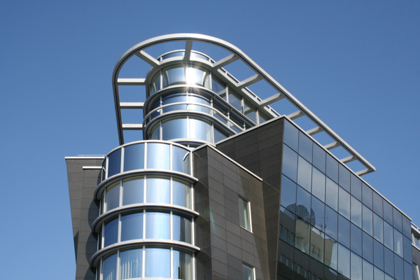 Modernes teilweise foliertes Gebäude mit großer Glasfassade und abgerundeten Fensterfronten, reflektierend im Sonnenlicht vor blauem Himmel.